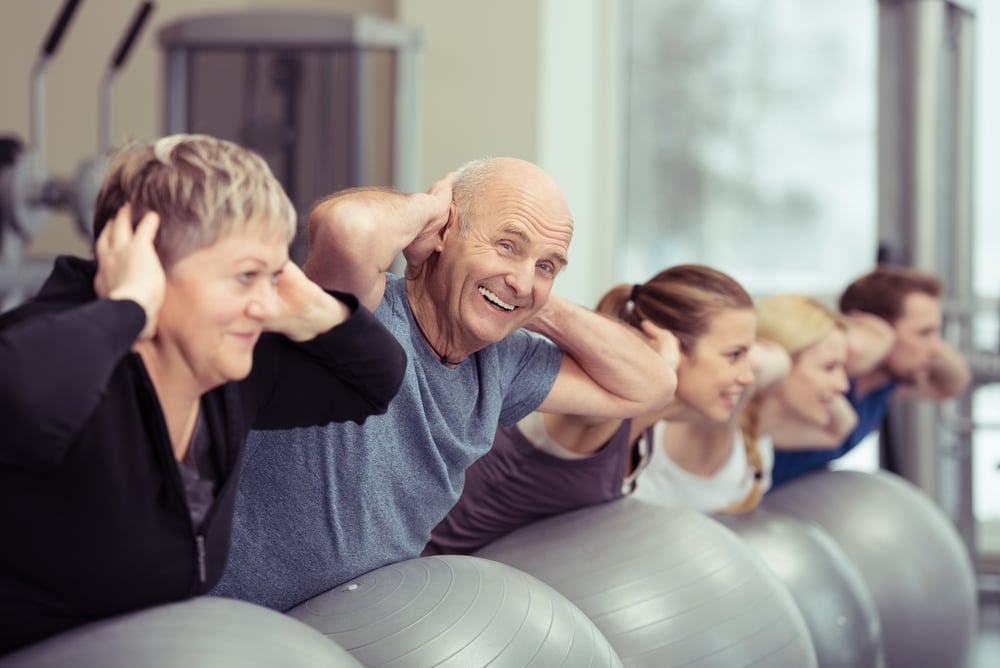 Elderly couple doing pilates class at the gym with a group of diverse younger people balancing on the gym ball with raised arms to tone their muscles in an active retirement concept