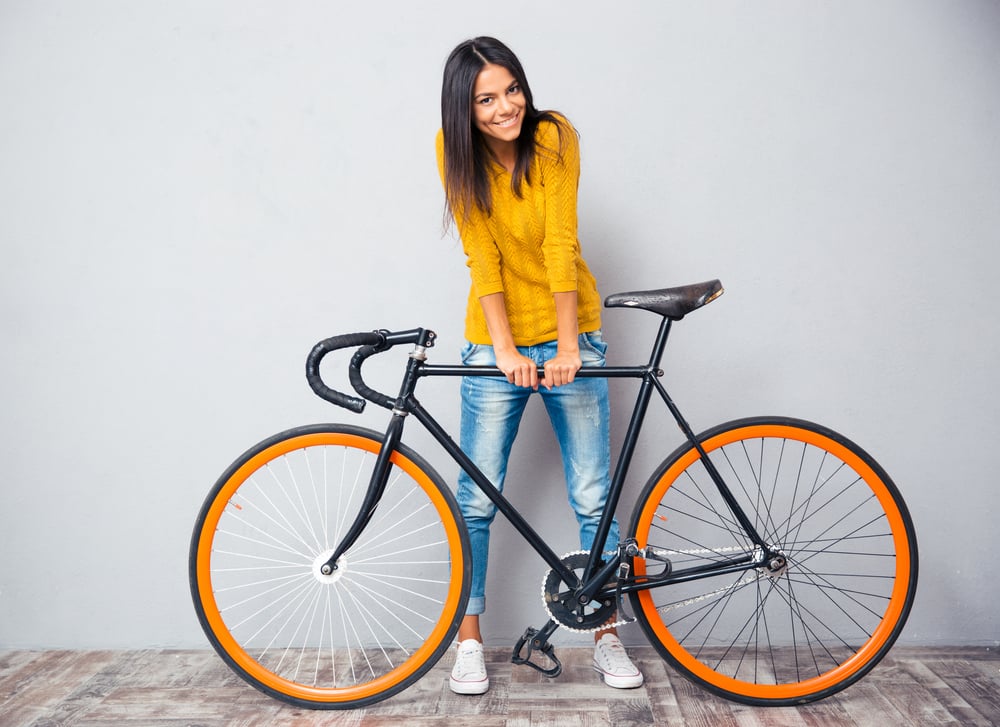 Full length portrait of a smiling woman standing near bicycle on gray background. Looking at camera