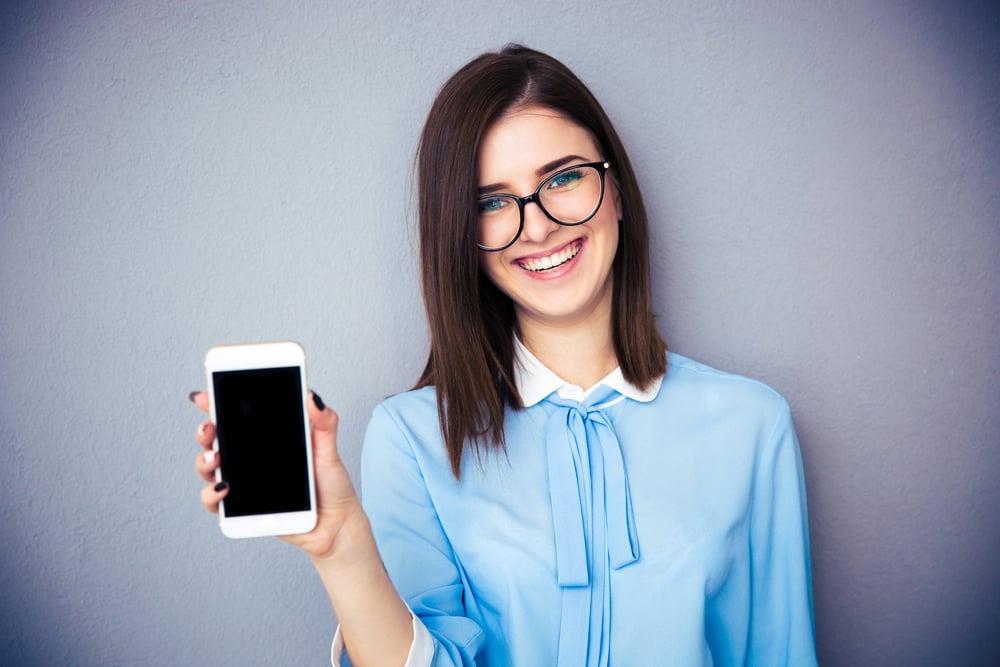 Smiling businesswoman showing blank smartphone screen over gray background. Wearing in blue shirt and glasses. Looking at camera.