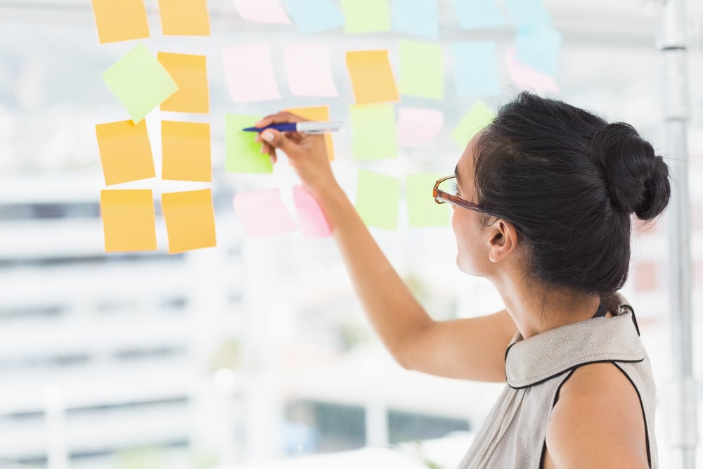 Smiling designer writing on sticky notes on window in creative office