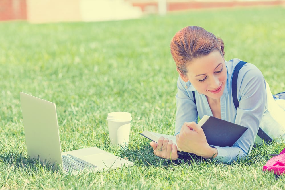 Student studying in park. Joyful happy young girl student sitting reading book outside on university campus or park. Education concept. Positive face expression