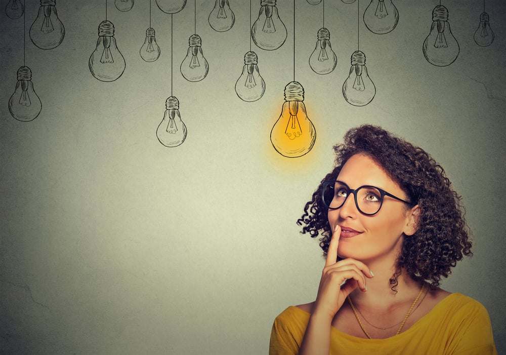 Thinking woman in glasses looking up with light idea bulb above head isolated on gray wall background-1