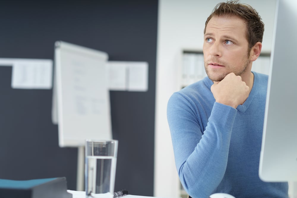 Thoughtful young businessman sitting at a desk in the office watching something to the left side of the frame with an engrossed expression